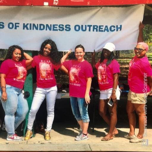 Volunteers in R.E.A.L. Women Ministry shirts standing under the Seeds of Kindness Outreach banner at a community event